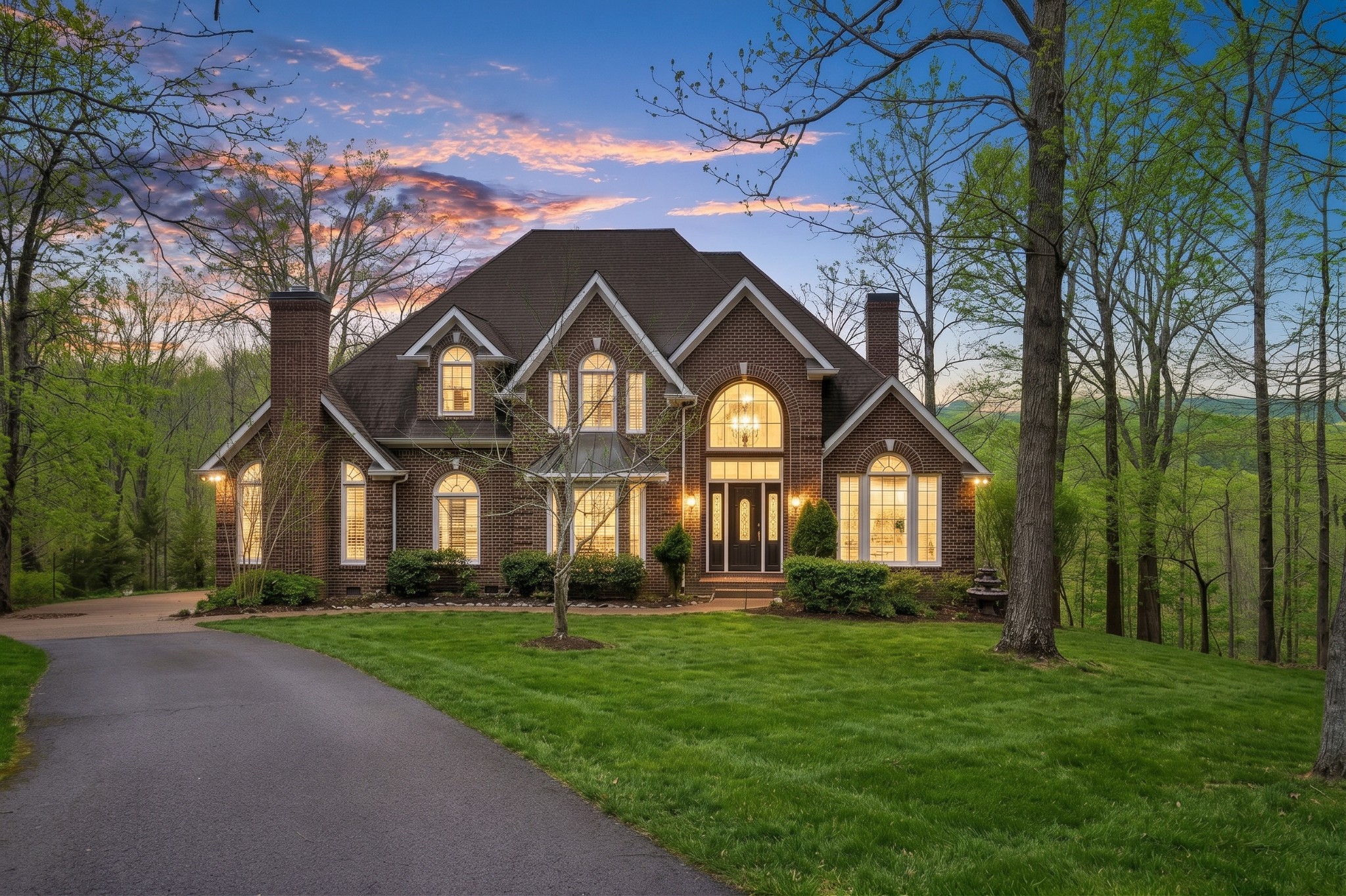 Brick two-story house with arched windows and warm interior lights, surrounded by tall trees at dusk.