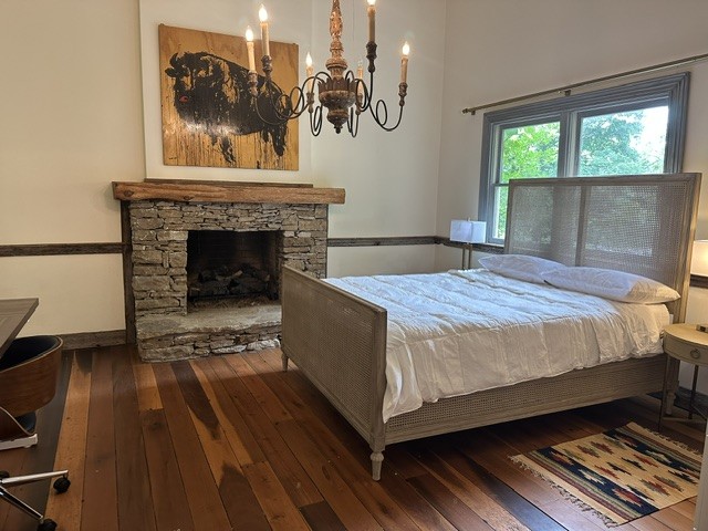 Bedroom with a stone fireplace and wooden mantel, an ornate iron chandelier, and a wicker-framed bed with white linens. A window shows greenery outside; hardwood floors and a small rug lie beside the bed.