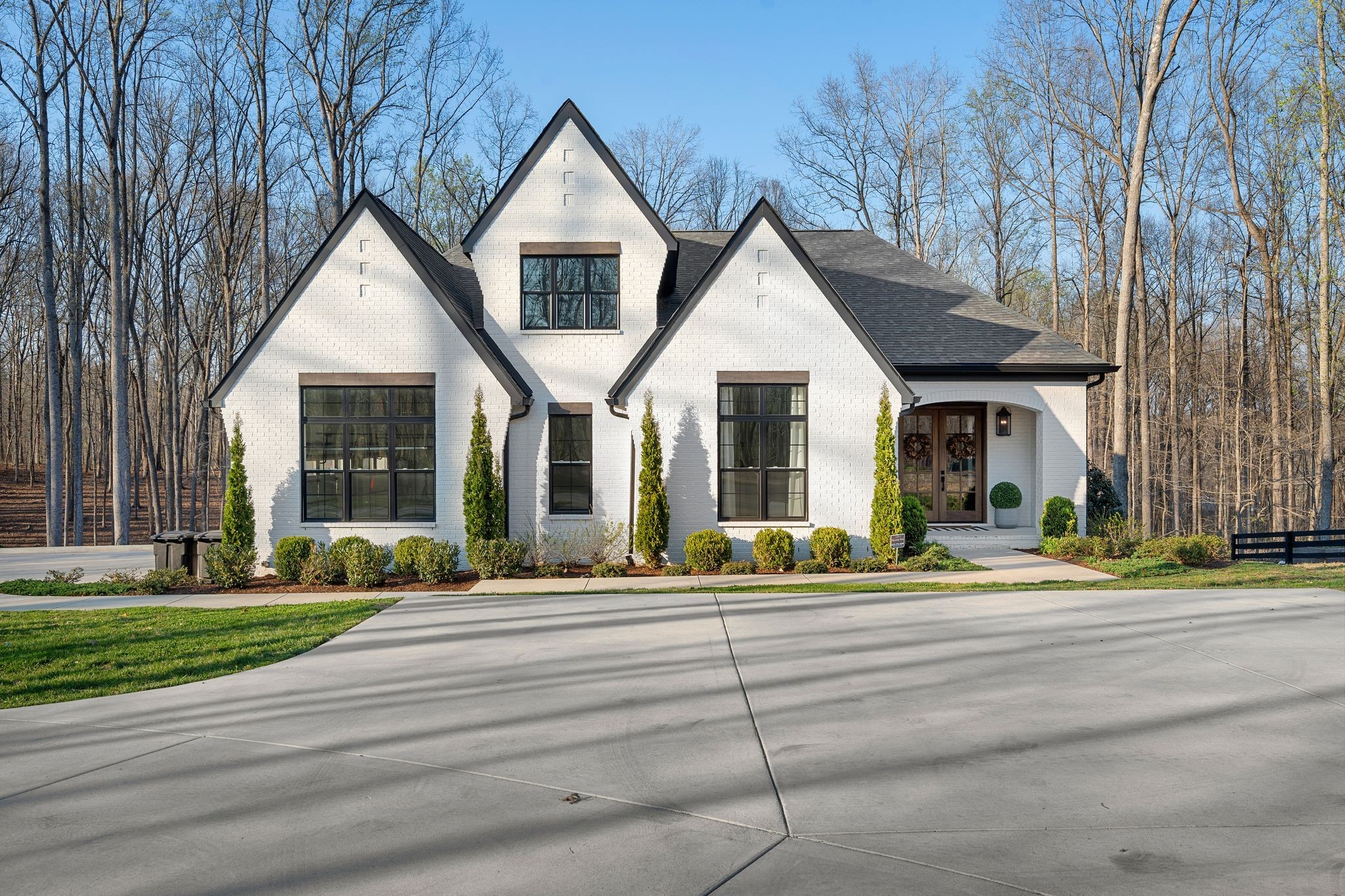 White brick house with three peaked gables, large black-framed windows, and a manicured line of shrubs along a concrete driveway.